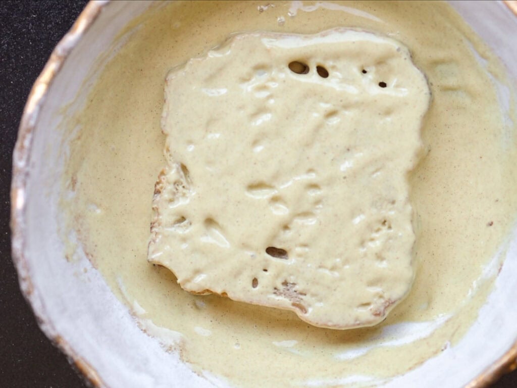 A slice of bread coated in a thick, pale yellow batter sits in a white bowl, ready to be cooked.